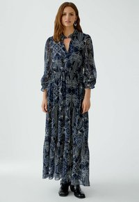 Langes navyblaues Kleid mit Paisleymuster, featuring einen Kragen, einem Frontschlitz und Puffärmeln. Fällt bis zu einem gestuften, gerafften Saum.
