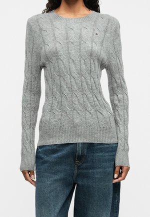 Kvinde iført en lysegrå kabelstrikket sweater med lange ærmer og blå denimjeans, stående mod en ensfarvet hvid baggrund.