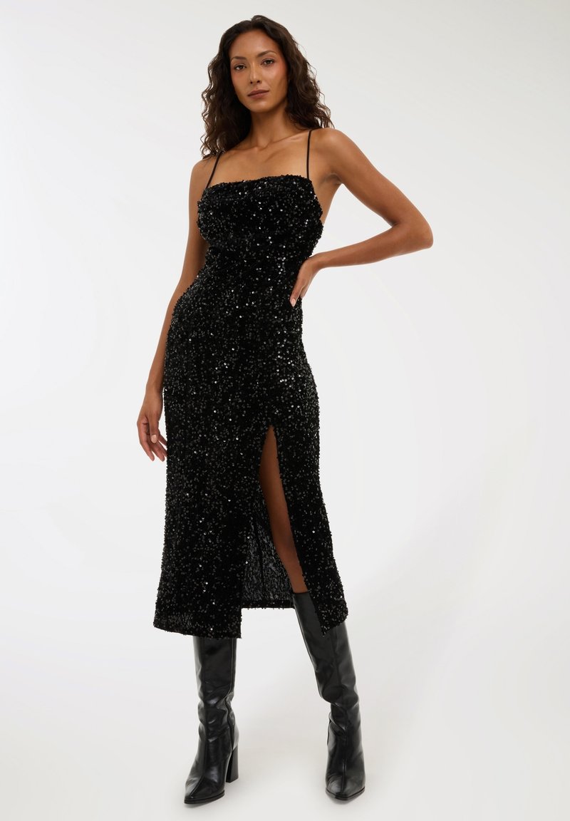 Kobieta w czarnej, błyszczącej sukience midi na ramiączkach, z rozcięciem na udzie, w zestawieniu z czarnymi, wysokimi booti na obcasie.