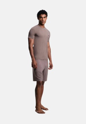 Uomo che indossa una camicia a maniche corte e pantaloncini abbinati in tonalità mauve smorzate, in piedi a piedi nudi su uno sfondo semplice e chiaro.