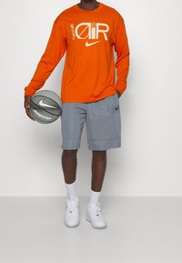 Orange long-sleeve Nike shirt with white logo, gray shorts, white socks, and Nike basketball in hand. Classic athletic wear style.