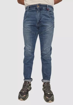 Uomo che indossa jeans blu con orli risvoltati e scarpe da ginnastica nere, in piedi davanti a uno sfondo chiaro semplice.