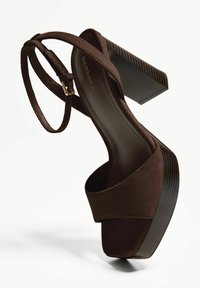 Sandali platform in suede marrone con una larga fascia, una sottile fascia alla caviglia e un tacco chunky testurizzato. Presentano un accento di fibbia dorata.