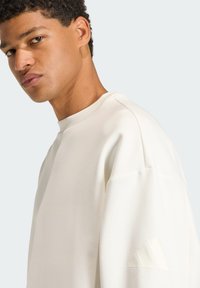 Cream-colored sweatshirt featuring a ribbed crew neck, dropped shoulders, and tonal Adidas logo on the left sleeve. Smooth texture.