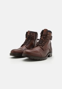 Bottes en cuir marron à la cheville avec un bout rond, un laçage à l'avant et une fermeture éclair sur le côté. Elles présentent un dessus texturé et une semelle en caoutchouc robuste.