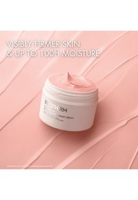 Hvit rund beholder med rosa kroppskrem med en jevn overflate. Etiketten har svart tekst på hvit bakgrunn, liggende på en rosa, teksturert bakgrunn.