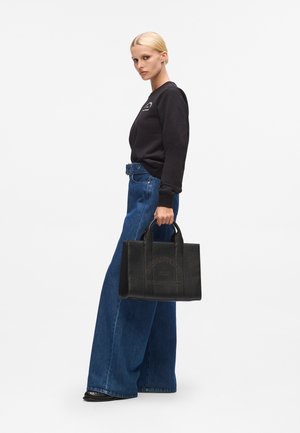 Blond kvinde iført sort langærmet top og brede denimjeans holder en sort læder Karl Lagerfeld taske ved sin side.