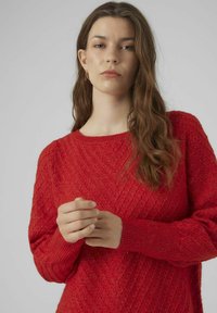 Felpa rossa con un motivo a coste diagonali e texture, scollatura rotonda e maniche lunghe con polsi aderenti. Il modello tiene le mani unite.