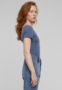 Modra majica s kratkimi rokavi iz mehkega materiala, prilegajoča se zasnova, nosena z visokim pasom svetlo modrih kavbojk. Gladka tekstura, minimalističen stil.