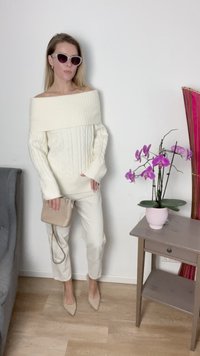 Suéter de malha em ponto de cabos creme com um design à mostra dos ombros, combinado com calças creme e saltos beije claros. Uma pequena bolsa beije e uma orquídea rosa ao lado.