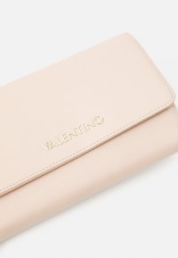 Valentino maksābe gaiši rozā teksturētā materiālā ar zelta logotipu. Iezīmējas ar locīšanas dizainu un gludu apdari.