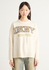 Kremfarget genser med strikkedesign, brede ribbestrikkede mansjetter og grafisk "DKNY NEW YORK" i mørk og lys brun skrift.