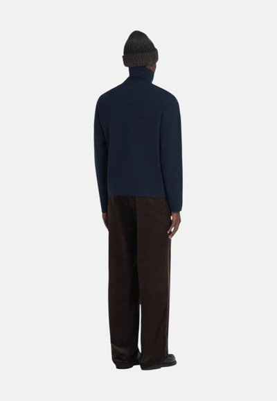 Pull à col roulé bleu marine en tissu lisse, associé à un pantalon en velours côtelé marron ample. Le modèle porte un bonnet en tricot foncé.