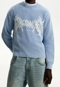 Ljusblå stickad tröja med ribbat mönster, med ett vitt grafiskt mönster i mitten och rundad halsringning. Matchad med ljusa denimjeans.