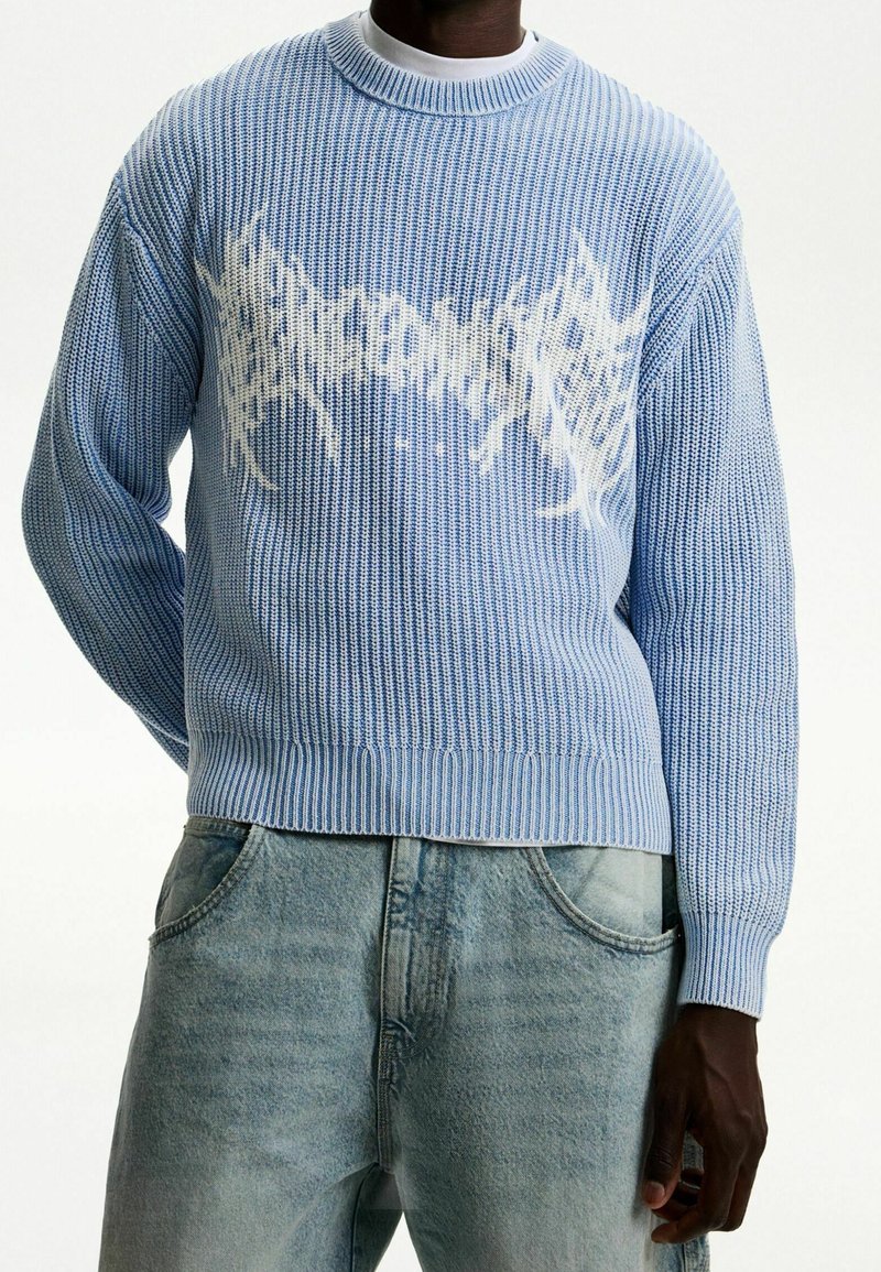 Ljusblå stickad tröja med ribbat mönster, med ett vitt grafiskt mönster i mitten och rundad halsringning. Matchad med ljusa denimjeans.