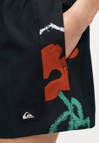 Κοντινό πλάνο μαύρων σορτς με το χέρι στην τσέπη, με αφηρημένο κόκκινο, πράσινο και λευκό μοτίβο και ένα μικρό λογότυπο Quiksilver κοντά στο τελείωμα.