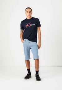 Tumši zili kokvilnas T-krekls ar baltu un sarkanu "Tommy Jeans" logotipu, apvienots ar gaiši zili džinsu īsajām biksēm un melniem sporta apaviem. Tīrs dizains, ikdienišķs izskats.