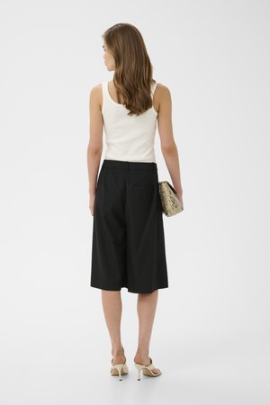 Kvinde med langt brunt hår iført en hvid ærmeløs top, sorte culottes med brede ben, beige hælede sandaler, der holder en clutch med slangeskind, vendt væk.