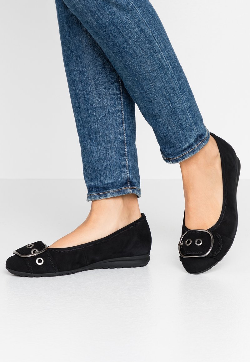 Personne portant un jean bleu slim et des chaussures plates noires avec une grande boucle décorative sur un fond clair.