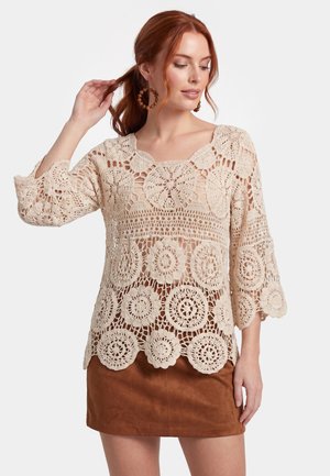 Rødhåret kvinde iført runde træbøjle-øreringe, beige hæklebluse med blomsterprint og brun ruskind mini-nederdel, poserer med hånden i håret.