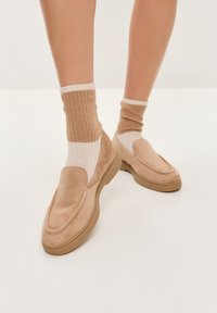 Bež slip-on cipele sa glatkom zamšinom i gumenim đonom, uparene s tamnosmešnim rebrastim čarapama sa kremastim prugama.