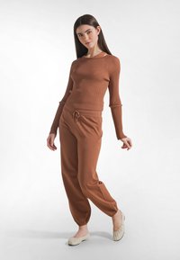 Mujer con top marrón de manga larga acanalado y pantalones a juego posando con zapatos planos beige sobre fondo blanco liso.