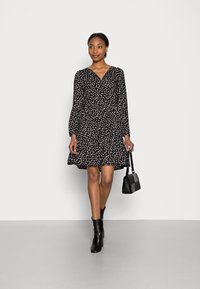 Vestido preto com decote em V, mangas longas e bainha evasê, apresentando um padrão abstrato branco. Combinado com uma pequena mala preta e botas de tornozelo.