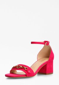 Sandal alte in suede rosa con cinturino alla caviglia, punta aperta e catena dorata decorativa sulla parte superiore. Tacco quadrato per maggiore stabilità.