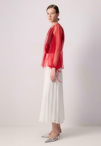 Transparente rote Overlay-Bluse mit elastischen Bündchen, kombiniert mit einem gefalteten weißen Midirock. Das Model trägt silberne spitze High Heels.