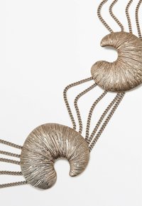 Collana in metallo con due pendenti curvi a forma di conchiglia in bronzo, caratterizzati da una superficie texturizzata e collegati da multiple catene, che mettono in evidenza un'intricata lavorazione.