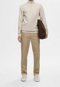 Sweatshirt beige clair à col zip, pantalons beiges et baskets crème ; tenant un sac marron. Texture lisse et coupe décontractée.