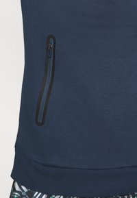 Close-up van een marineblauwe kledingstuk met een verticale ritssluitingzak omlijnd in zwart nabij de taille, gedragen over een patroonbroek.