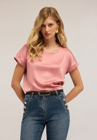 Top roz din satin cu mâneci scurte răsucite, asortat cu blugi închiși la culoare, cu talie înaltă, purtați cu o curea neagră și accente cu nasturi.