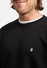 Sort sweatshirt lavet af bomuld med ribbet hals. Har et lille hvidt logo på brystet. Glat tekstur, afslappet pasform, enkelt design.