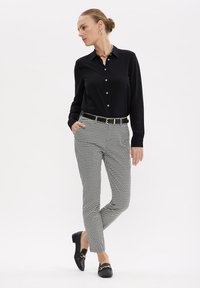 Camicia nera a maniche lunghe con bottoni, pantaloni neri e bianchi a motivi, cintura con dettagli dorati e mocassini neri con dettagli in metallo.