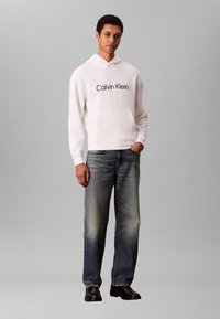 Jauns vīrietis baltā Calvin Klein sporta krekliņā, izbalējās zilās džinsās un melnās kurpēs, stāvot ar vienu roku kabatā uz pelēka fona.