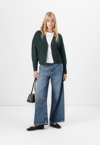 Kvinne som står mot hvit bakgrunn iført mørkegrønn knappet cardigan, hvit skjorte, vide blå jeans, svarte leiligheter, holder en liten brun håndveske.