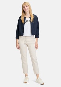 Veste de bombardier marine avec des poignets élastiques, T-shirt blanc à imprimé graphique, pantalon beige ajusté et baskets légères. Design simple et couleurs neutres.