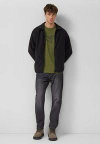 Zwarte fleecejas, groen T-shirt met lijnpatroon, donkere spijkerbroek en bruine laarzen. Losse pasvorm met ritssluiting en lange mouwen.