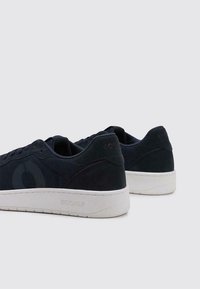 Baskets bleu foncé avec un tissu texturé et une finition en daim, dotées d'une semelle en caoutchouc blanche et d'un logo de marque sur le talon et sur le côté.