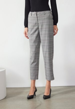 Femme portant un pantalon gris à carreaux longueur cheville avec un haut noir et des chaussures à talons noirs, debout sur un sol lisse contre un mur blanc.