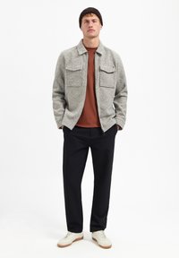 Lichtgrijze textuurjacket met twee voorkevels, roestkleurig shirt eronder, zwarte broek en witte sneakers, van voren bekeken.