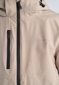 Beżowa kurtka z przodu z czarnymi, wodoodpornymi zamkami błyskawicznymi i kapturem ze sznurkiem, z subtelnym logo "LINDBERGH" na piersi.