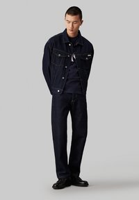 Veste en denim foncé avec une fermeture à boutons et deux poches poitrine, associée à un pantalon en denim foncé assorti et des chaussures noires. Texture lisse.