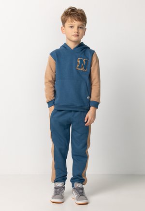 Jongen die staat, gekleed in een blauwe en tan hoodie met 'N' patch, bijpassende broek met tan zijstrepen, en grijze sneakers.