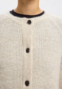 Beige stickad cardigan med ett texturerat tyg, med mörka knappar, rund halsringning och raglanärmar.