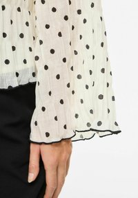 Biała bluzka w czarne kropki, przezroczysta fakturowana tkanina, mankiety z falbankami wykończone czarną lamówką, dopasowana w talii.