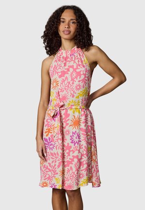 Kvinde med krøllet hår iført en ærmeløs, lyserød blomsterkjole med orange, gule og lilla mønstre, stående med den ene hånd på hoften.
