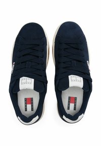 Sneakers in suede blu navy con lacci piatti, suole in gomma e etichette in tessuto marchiate. Presentano un design minimalista e accenti bianchi a contrasto.
