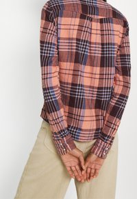 Chemise à carreaux aux couleurs orange, marron et bleu, dotée de manches longues et de poignets boutonnés, présentant une coupe décontractée et un tissu texturé.